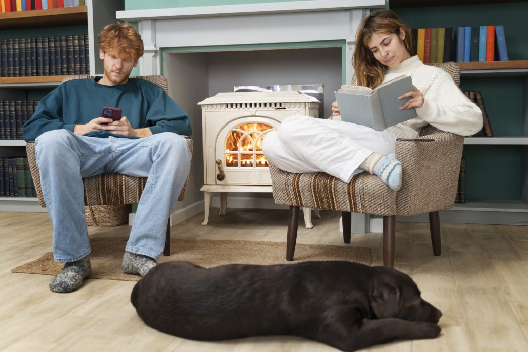 Couple relaxing indoors by a lit fireplace, with one person reading a book, the other using a smartphone, and a dog resting on the floor.